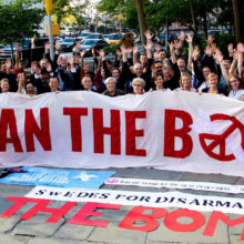 Joukko ydinasekieltoa kannattavia aktivisteja hurraa ja pitelee valkoista banderollia, jossa lukee punaisin kirjaimin "Ban the Bomb"